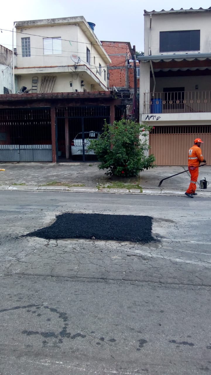 Buraco já foi tapado na rua Maria Luiza do Val Penteado. Trabalhador com macacão amarelo está na calçada com uma ferramenta na mão e um balde manchado de preto no chão. Ao fundo, sobrados simples na rua.